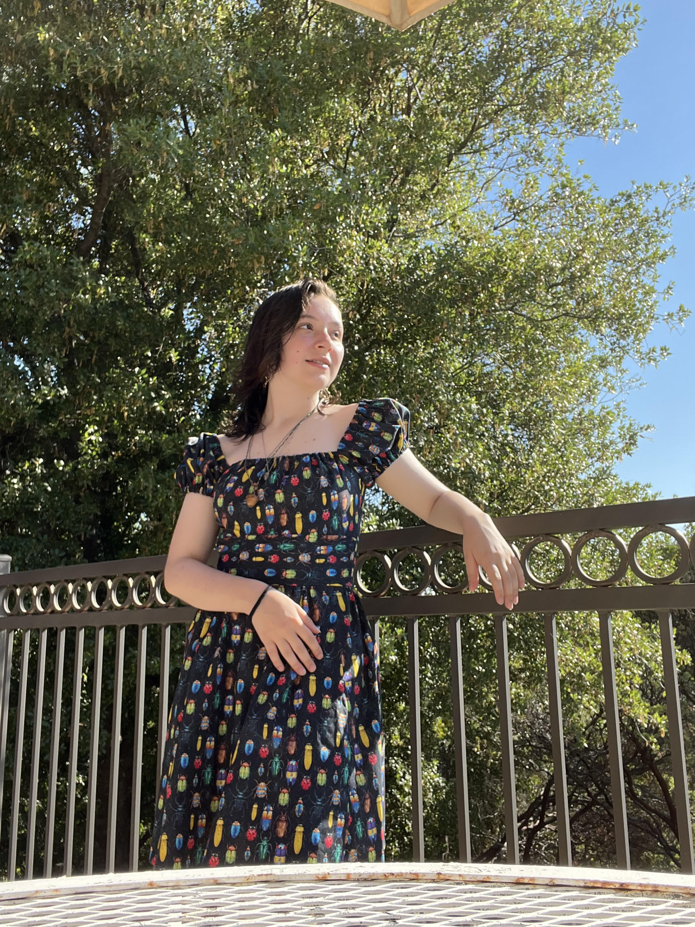 Mia stands on a balcony in a dress with a pattern of bugs on it