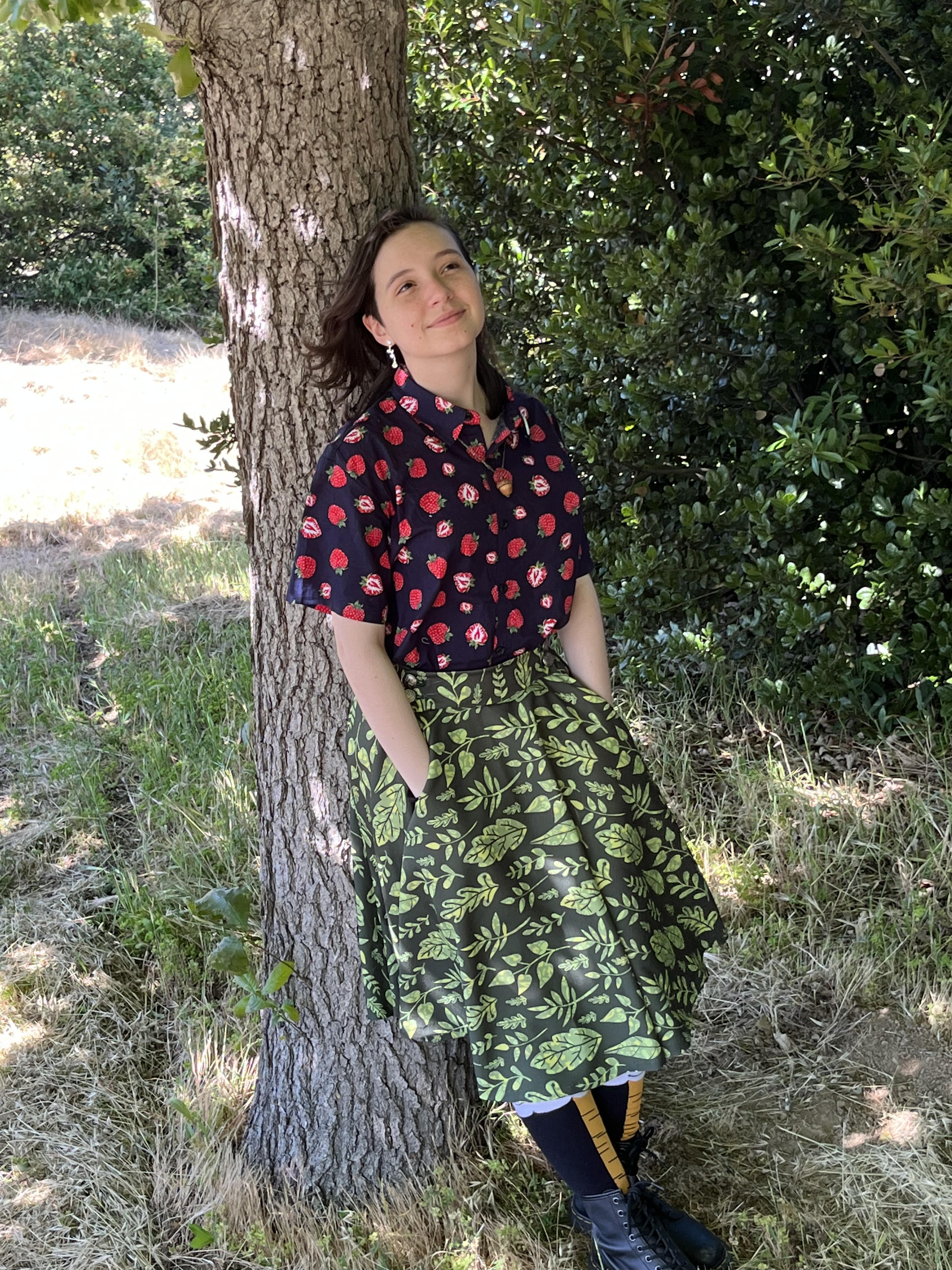 Mia leans on a tree in a shirt with strwberries and a skirt with green leaf patterns