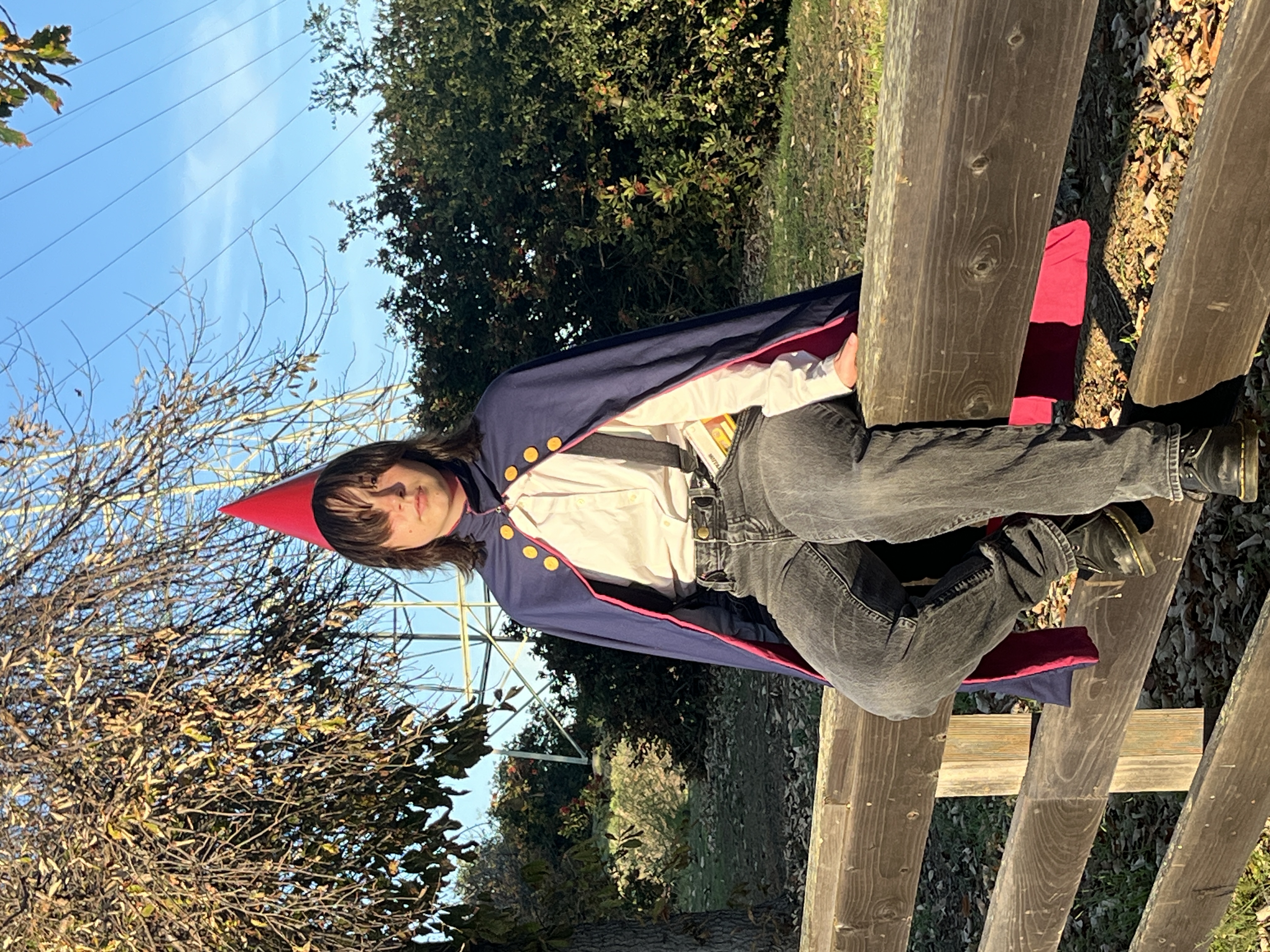 Mia dressed as Wirt from Over the Garden Wall sitting on a fence in front of woods