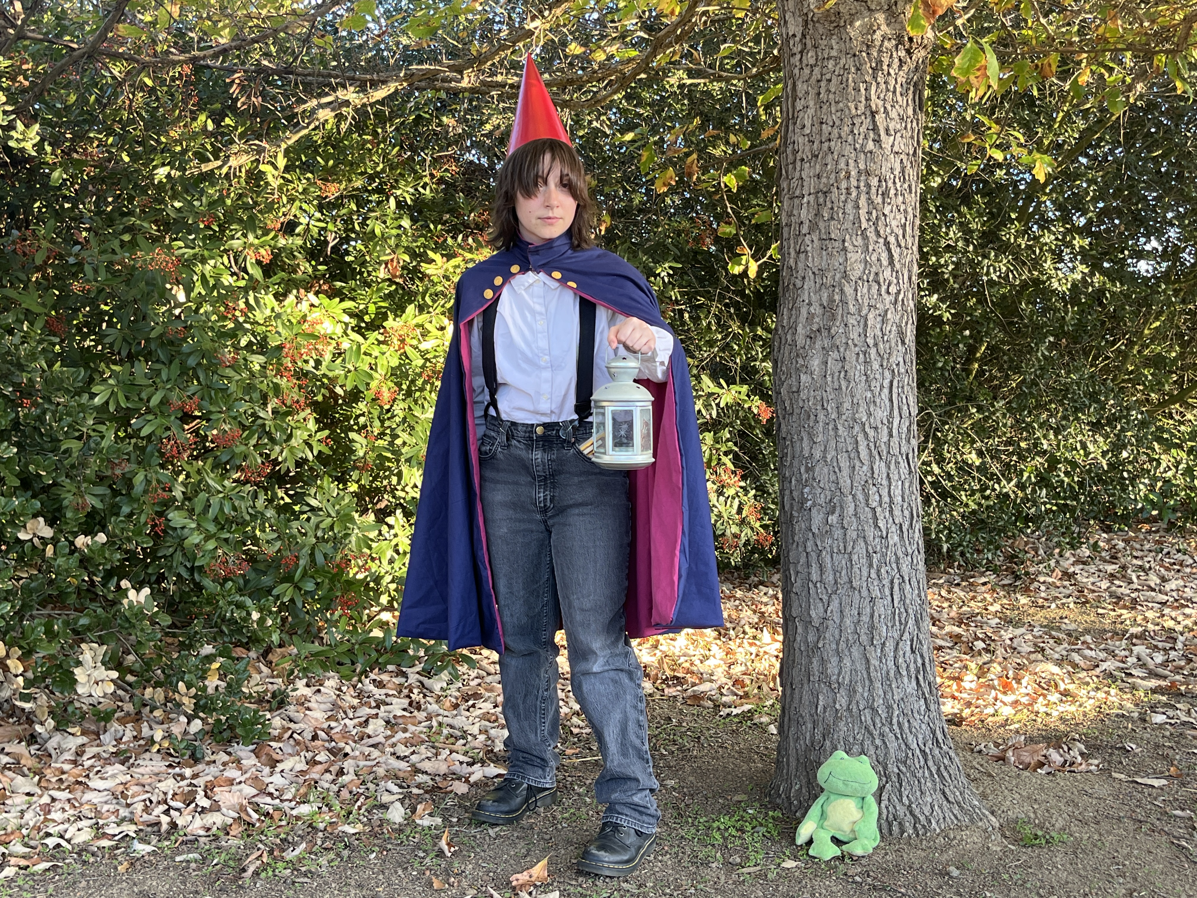 Mia dressed as Wirt from Over the Garden Wall holding a lantern in front of woods